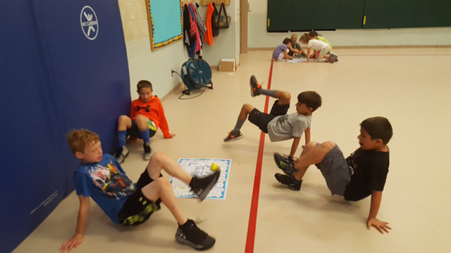 Students surrounding miniature mat on gym floor playing Fitness Skillastics